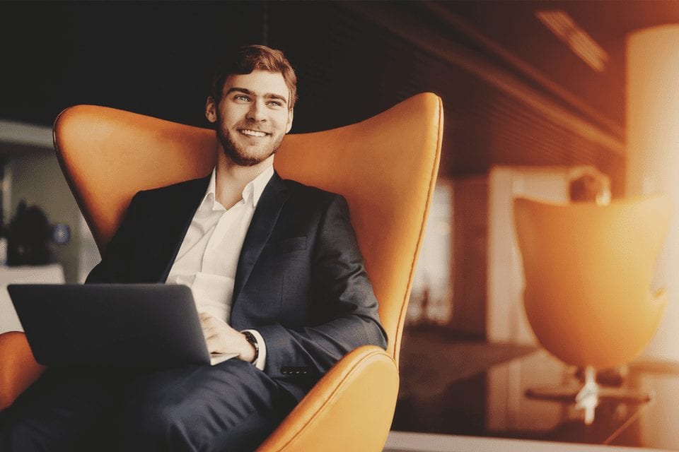 an image of a man sitting in a chair with a laptop on his lap. He is looking away and smiling.